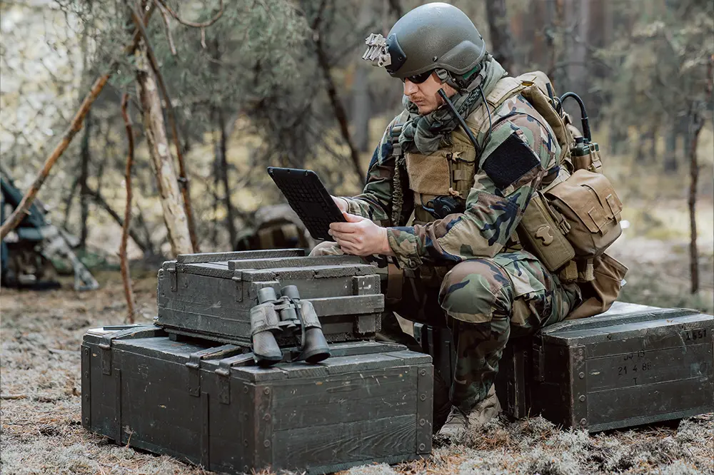 Soldat mit Tablet sitzt auf Munitionskisten im Wald