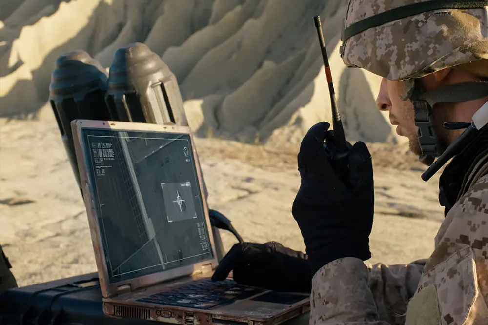Soldat mit Funkgerät arbeitet in der Wüste an einem robusten Laptop