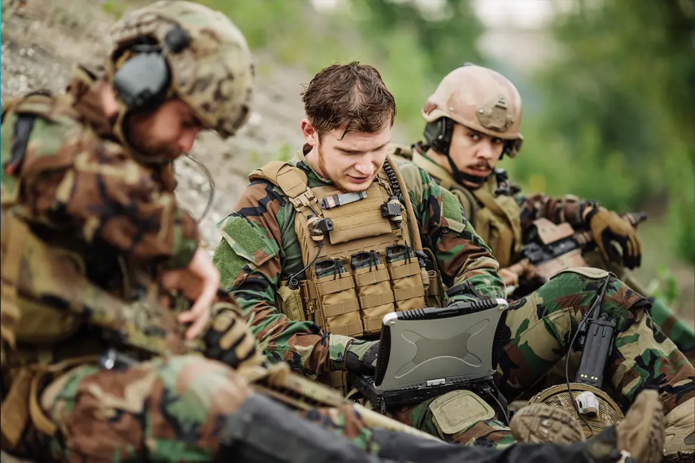 Soldaten im Gelände arbeiten gemeinsam an einem robusten Laptop