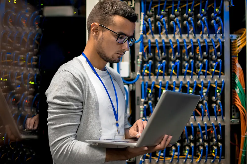 IT-Techniker mit Laptop vor einem verkabelten Netzwerkschrank