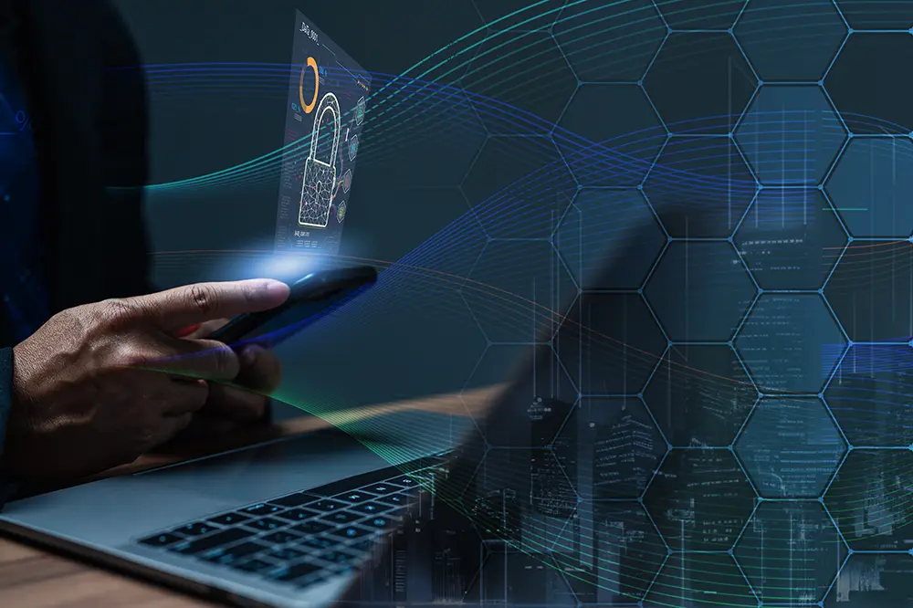 Hand mit Smartphone vor einem Laptop mit eingeblendeter Sicherheitsgrafik